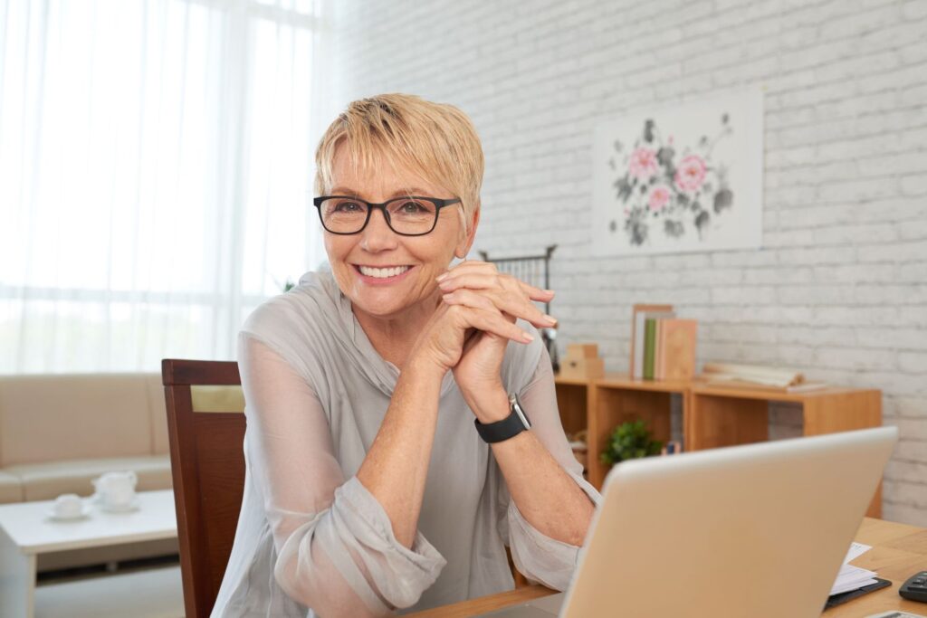 Comment gérer les bouffées de chaleur au bureau ? 4 Une femme ayant la cinquantaine regarde l'objectif souriante elle porte des lunette elle est installée devant son ordinateur à son bureau pour pour suggérer comment gérer les bouffées de chaleur au bureau.
