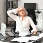 Une femme ayant la cinquantaine se tient le main sur le front et exprime l'impression d'avoir chaud pour suggérer comment gérer les bouffées de chaleur au bureau.