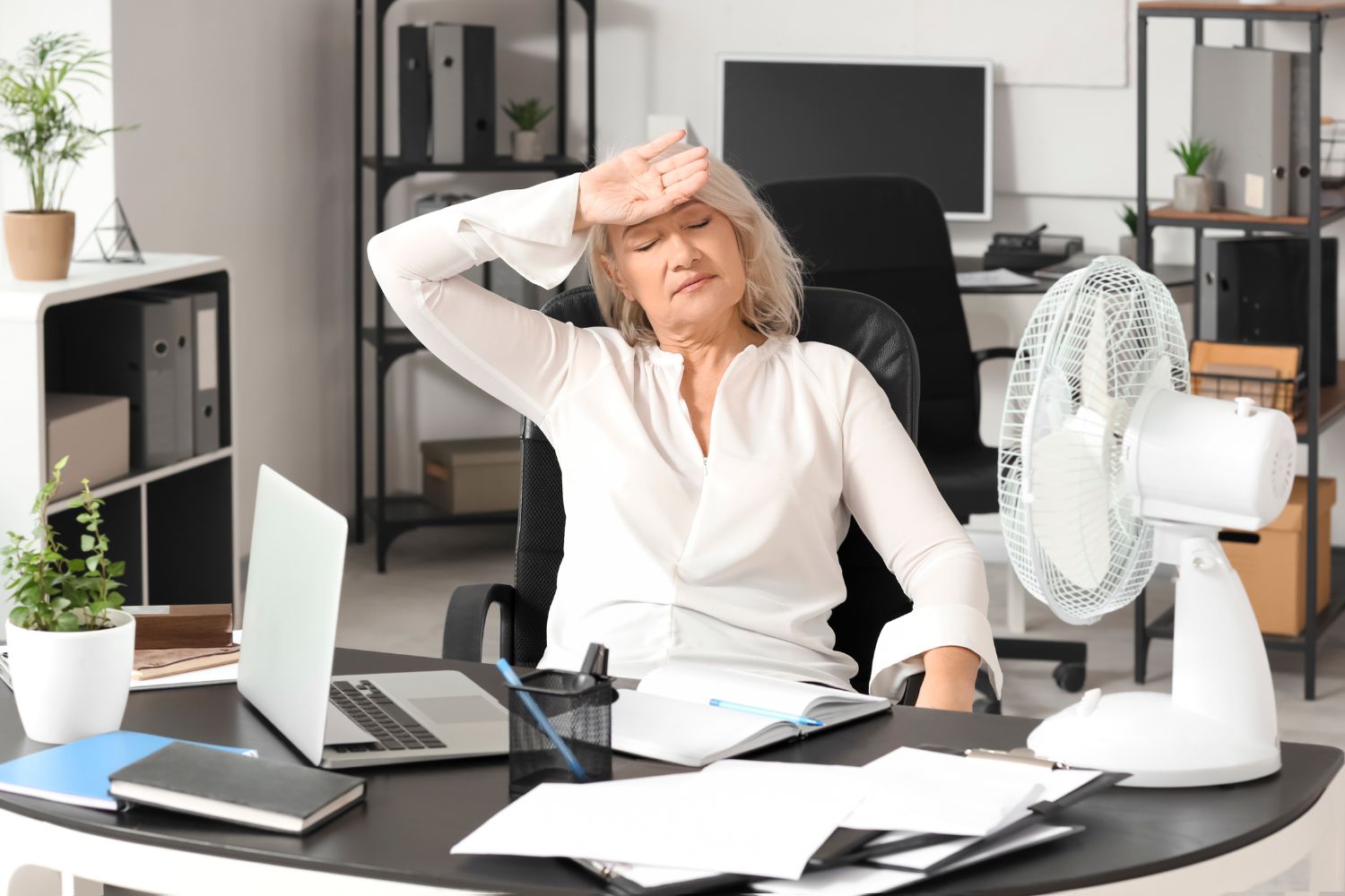 Une femme ayant la cinquantaine se tient le main sur le front et exprime l'impression d'avoir chaud pour suggérer comment gérer les bouffées de chaleur au bureau.