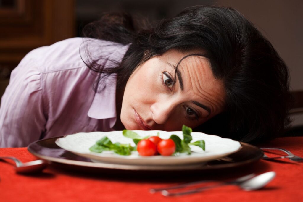 Perte de poids à la ménopause : stratégie douce et durable 2 une femme pose la tete sur la table l'air déprimé devant une assiette blanche qui contient 3 tomates cerises pour l'article perte de poids à la ménopause