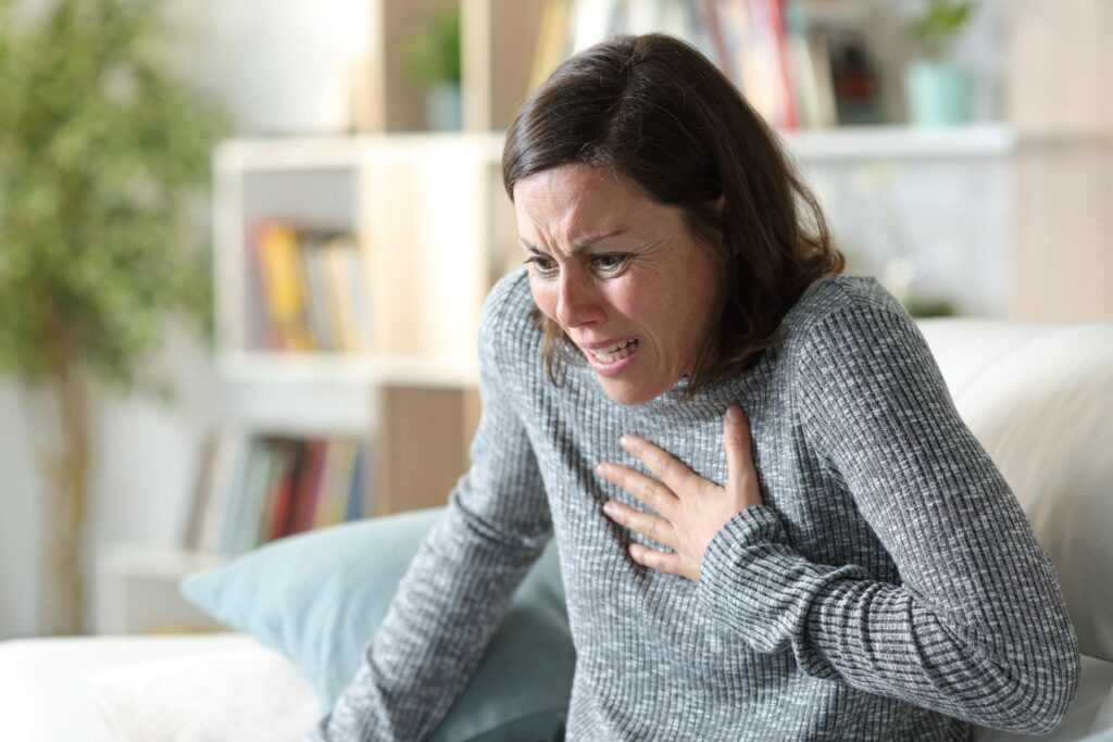 une femme cheveux brun porte un pull gris se tient la main sur la poitrine avec le visage crispé