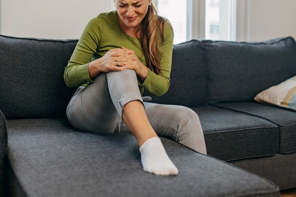 Une femme assise sur son canapé a le pied posé dessus et se tient le genou avec une expression de douleur sur le visage pour illustrer des douleurs articulaires à la ménopause