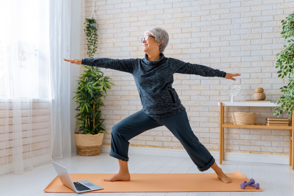 Une femme en tenue foncée de sport avec les cheveux gris porte des lunette et fait une posture de yoga pour illustrer l'article ménopause et sport