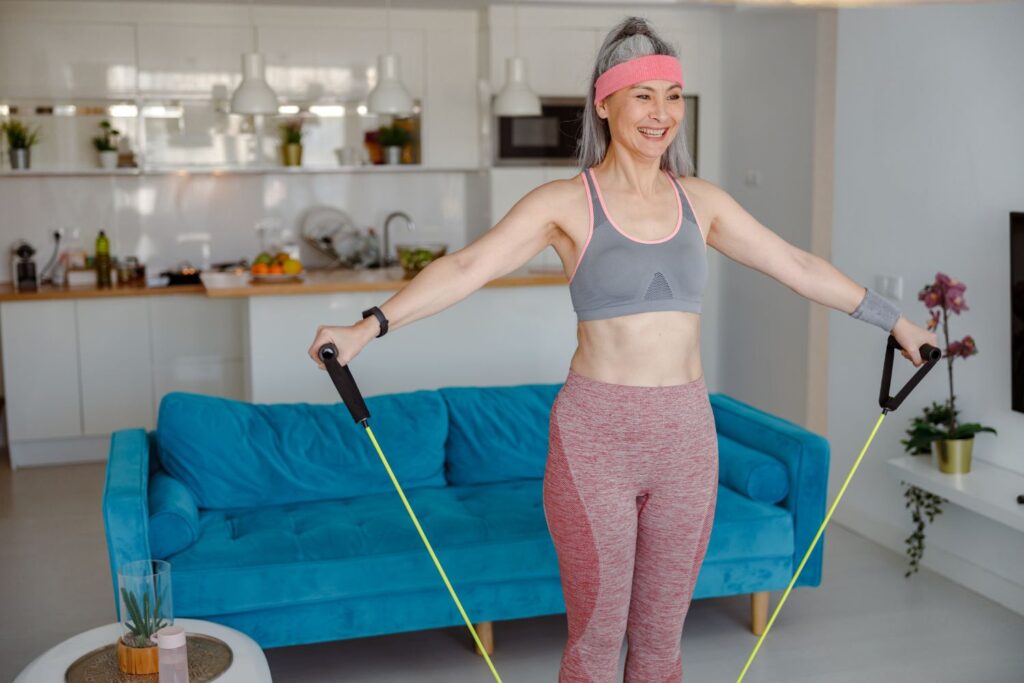 Une femme cheveux gris en tenue de sport rose et gris avec un bandeau rose sur le front fait su sport avec un élastique sous les pieds bras tendus pour illustrer l'article ménopause et sport