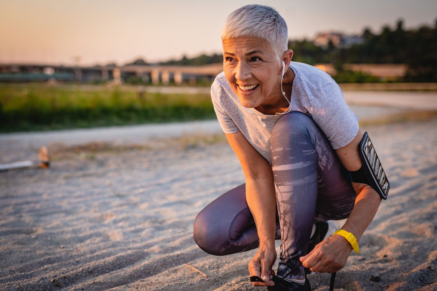 Une femme avec les cheveux courts et gris attache les lacets des ses baskets en tenue de sport a l'extérieur en soirée pour illustrer l'article ménopause et sport