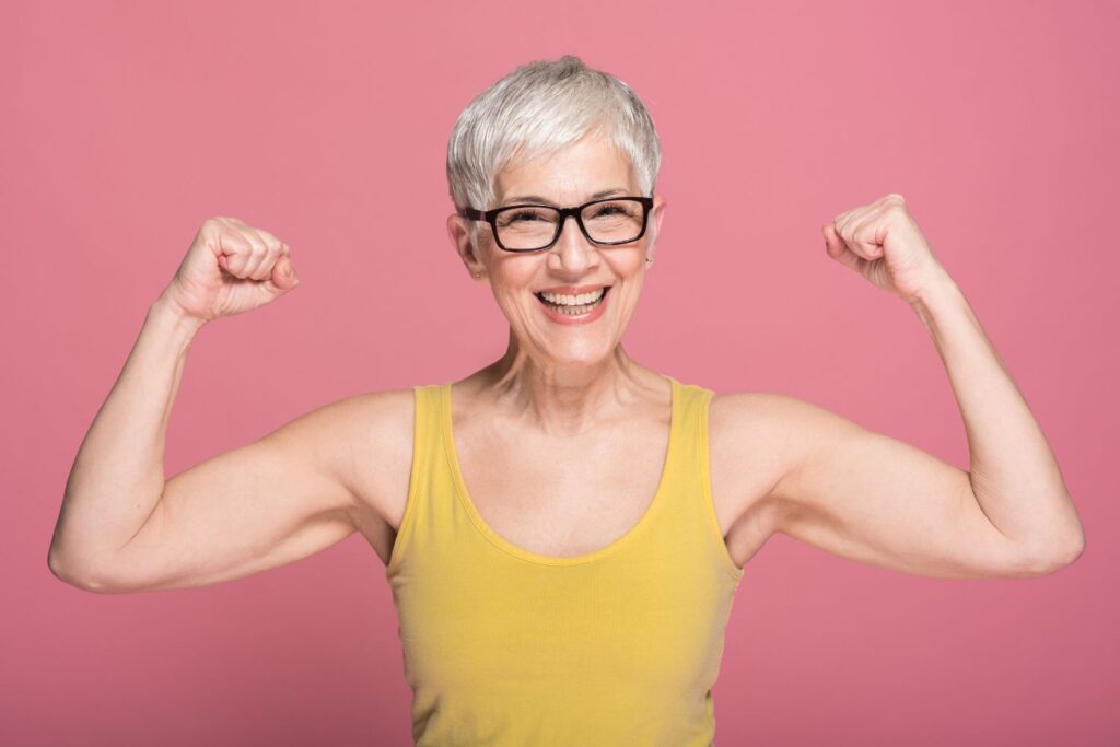 Une femme avec les cheveux courts et gris qui porte un haut jaune sur fond rose contracte ses biceps pour illustrer l'article ménopause et sport