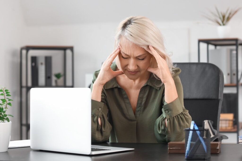Une femme la cinquantaine blonde devant son ordinateur sur son bureau se tient la tete entre ses mains pour illustrer l'article comment parler de la ménopause au travail