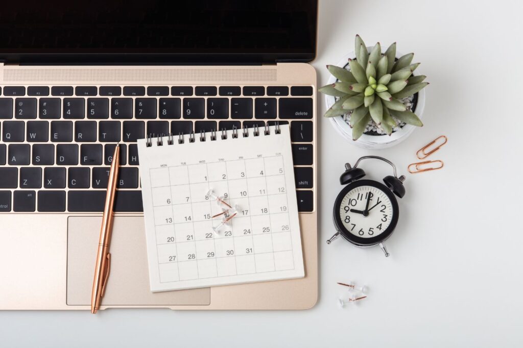 Un ordinateur portable, un style, un carnet calendrier et un réveil posés sur un support blanc pour illustrer l'article comment parler de la ménopause au travail