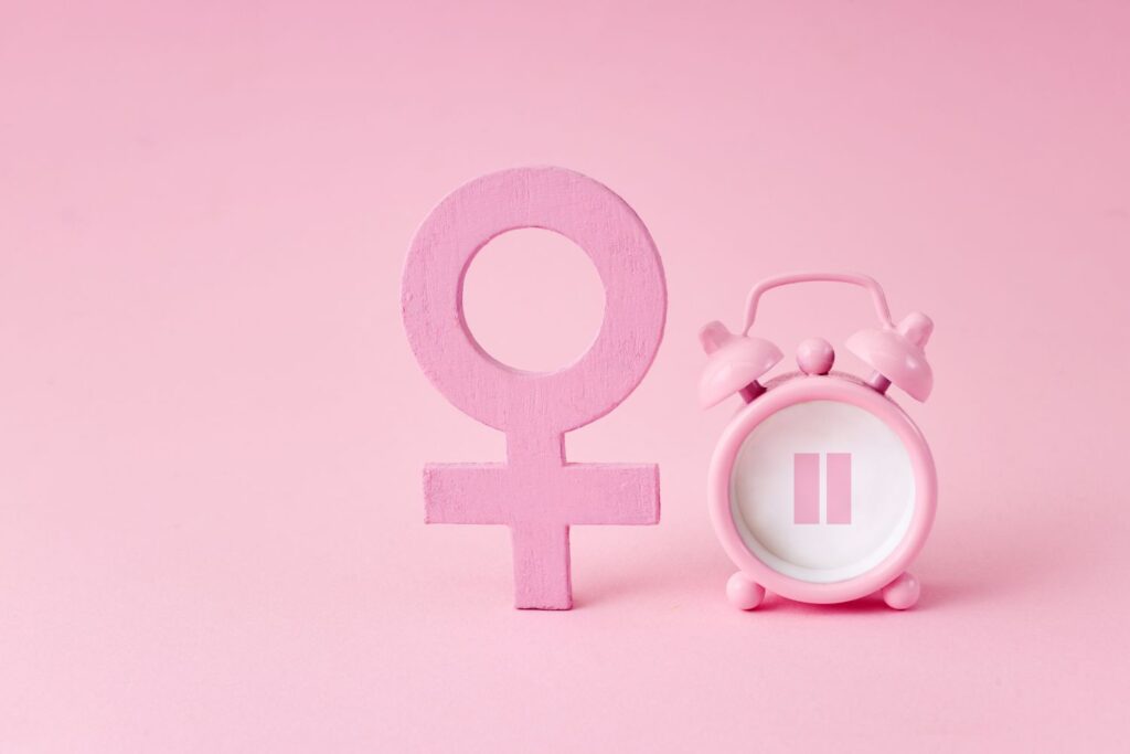 Le symbole physique du féminin à coté d'un réveil matin indiquant le signe pause à l'intérieur sur un fond rose pour illustrer l'article comment savoir si on est ménopausée quand on a un stérilet