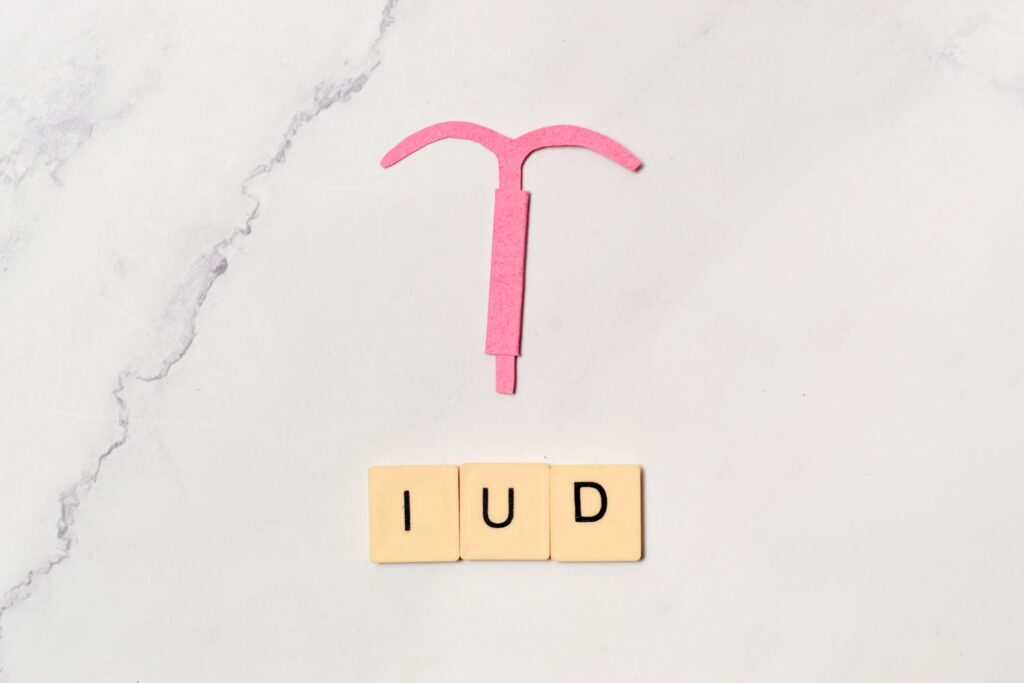 Une découpe de papier rose en forme de stérilet posée sur un fond gris marbré avec les lettre IUD en dessous pour illustrer l'article comment savoir si on est ménopausée quand on a un stérilet