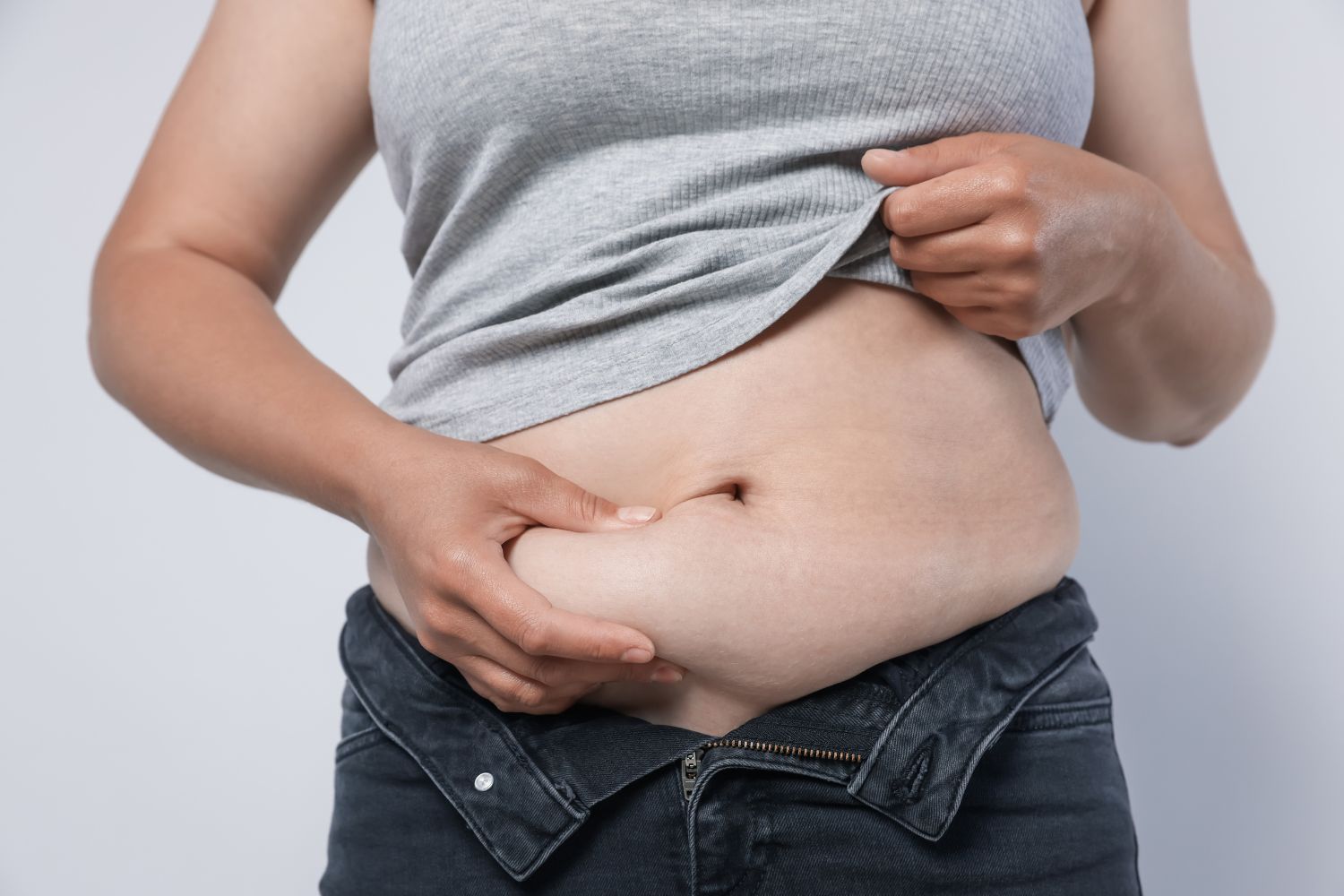 Une femme se tient debout de face lève son haut et pince la graisse sur son ventre à la ménopause